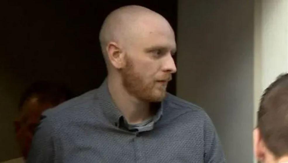 A man in his 30s with a bald head and beard is led out of court by security guards