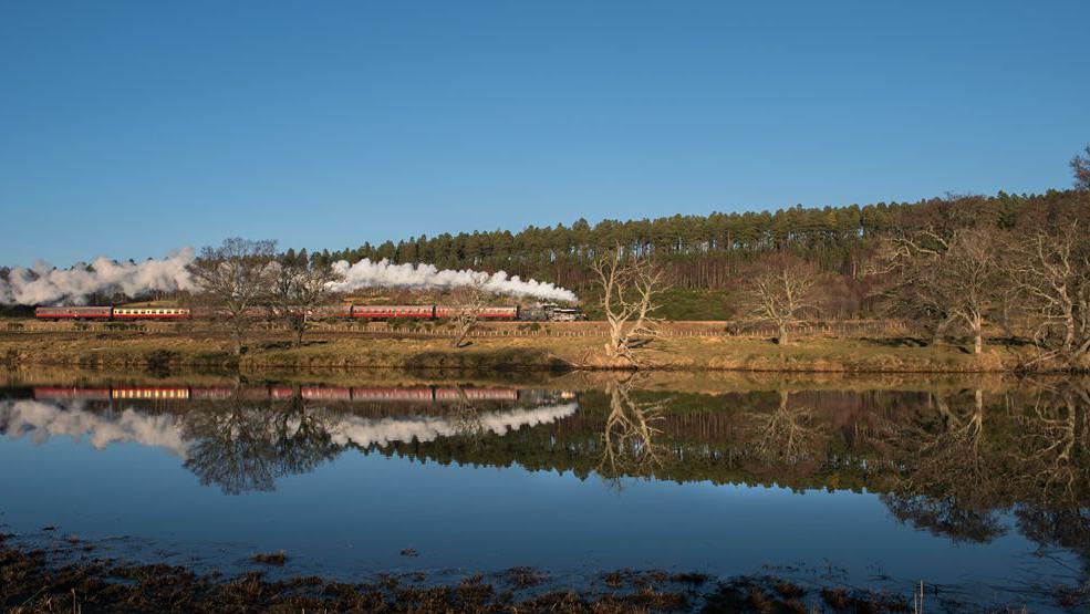 A black locomotive trailing a plume of smoke as it pulls carriages on a bright sunny day. The train is reflected in the still waters of a loch.