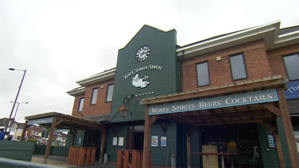 External shot of a brick-built pub with a large green panel featuring a clock and The Clumsy Swan pub sign