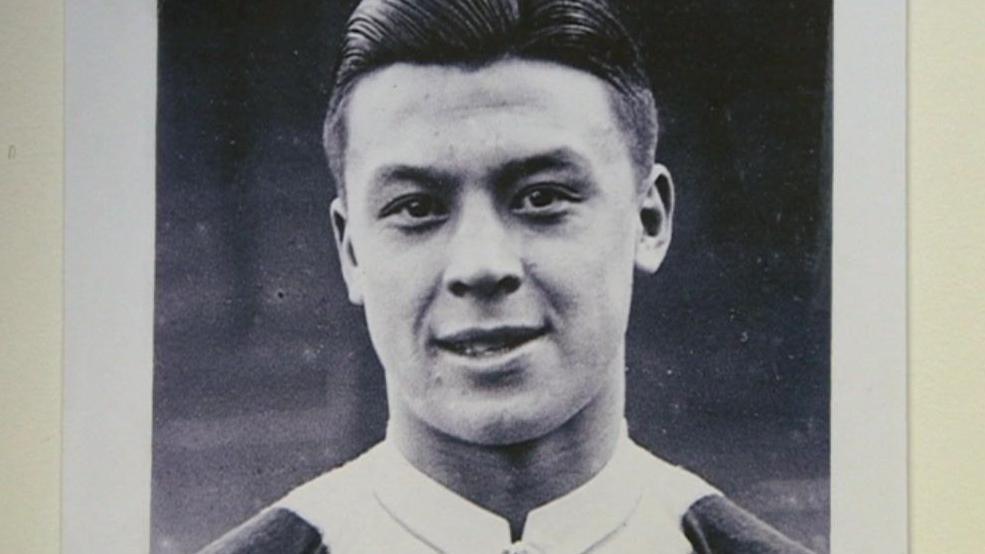 A black and white photo of Frank Soo, which shows him in his football kit with black, slicked-back hair and smiling for the camera.