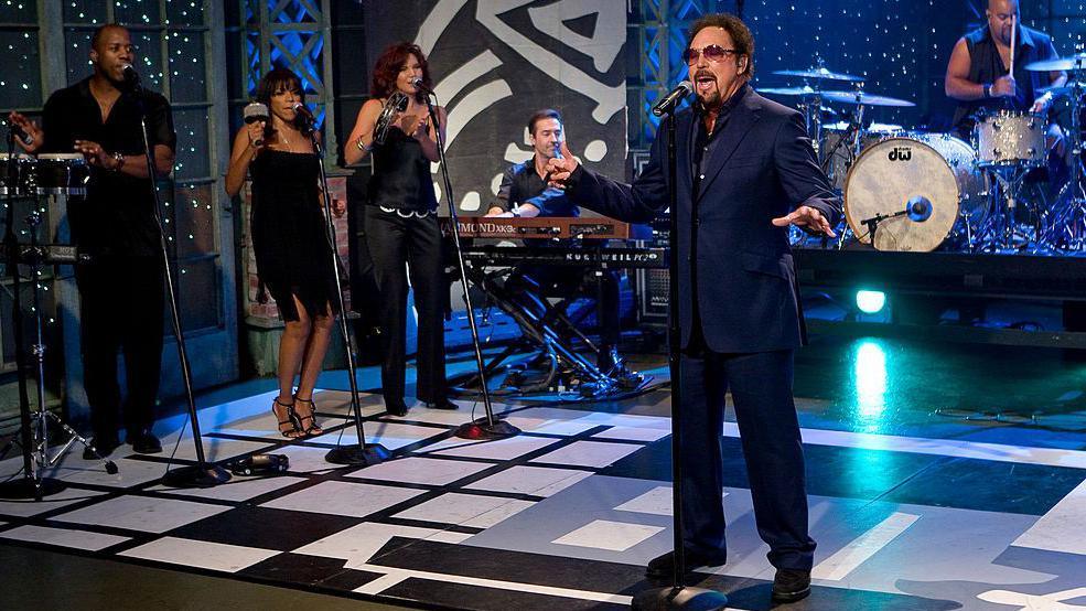 Tom Jones sings on stage with a five piece band including backing singers with percussion instruments, a drummer and a keyboard player. He is wearing a navy blue suit, aviator sunglasses with pink lenses and his hair and goatee beard are dyed brown.