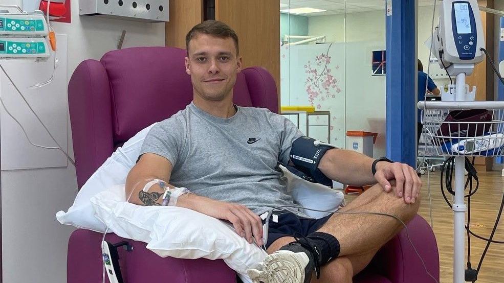 Liam Landers smiling at the camera while sitting in a purple chair on a hospital ward. He has tubes going into his arms, as he has chemotherapy at hospital. 