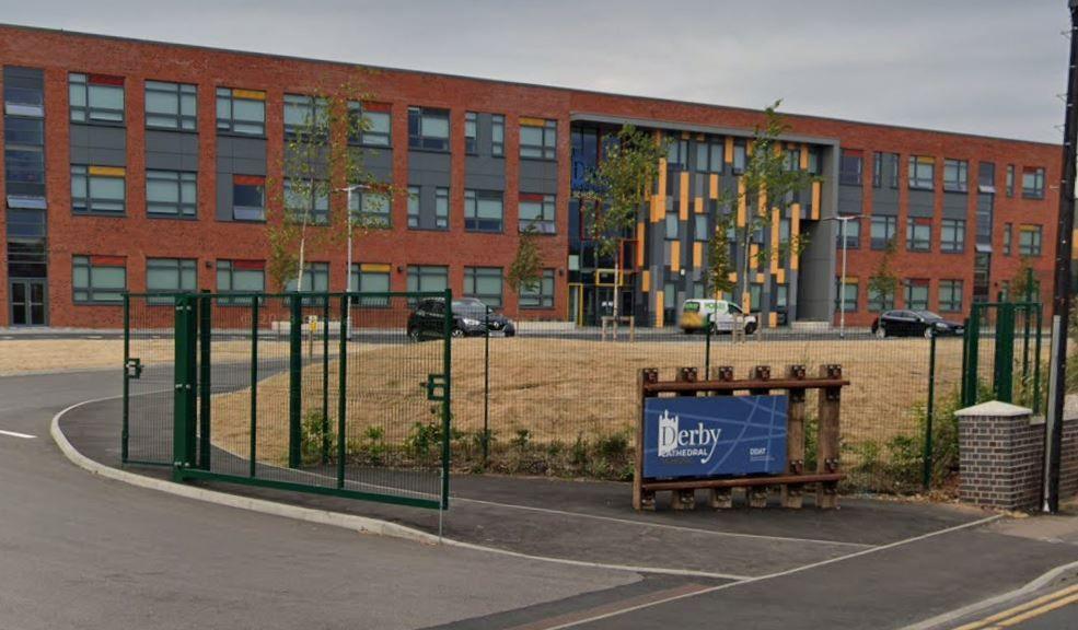 Image of a red-brick school with grey window frames and lawns of yellow grass surrounded by a racing green-coloured fence.