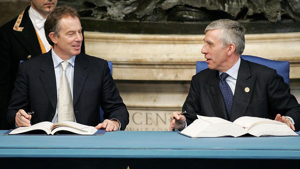 Tony Blair and Jack Straw sit at a table with large books in front of them and they are holding pens.