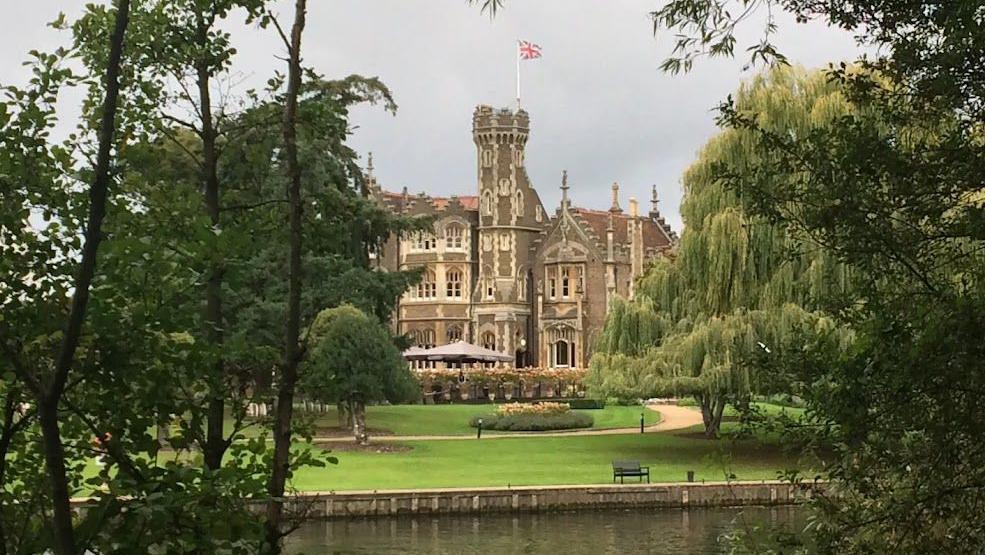 A brown stone turreted building with lighter stone detailing, ornate windows and two wide umbrellas outside is visible through a clearing between trees, facing on to a body of water and mowed lawns with flower beds . A bench and a bin are situated near the calm looking water.