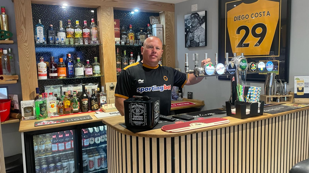 Glenn is stood in his bar. He is standing behind a wooden bar with panelling on the front which follows its curved shape. There are beers on draught and shelves full of spirits and drink behind him. There is also small fridge filled with beer behind him. To the right, a Wolves shirt of Diego Costa is hanging on the wall.