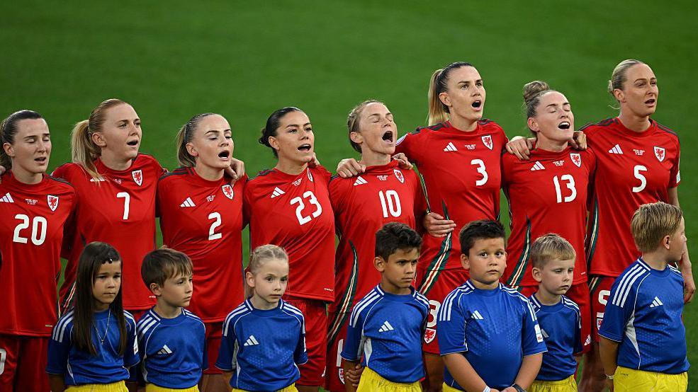 Eight Wales players, in red shirts, line up to sing the national anthem. In front of them are seven mascots wearing blue tops and white shorts
