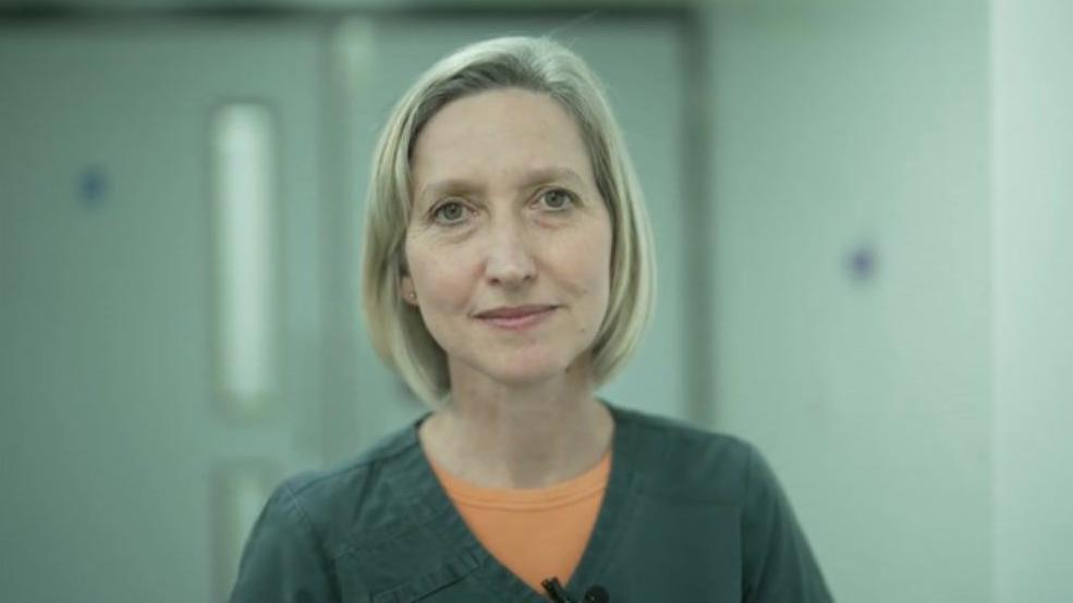 A woman with chin length blonde hair looks towards the camera. She is wearing green medical scrubs with a light orange top underneath