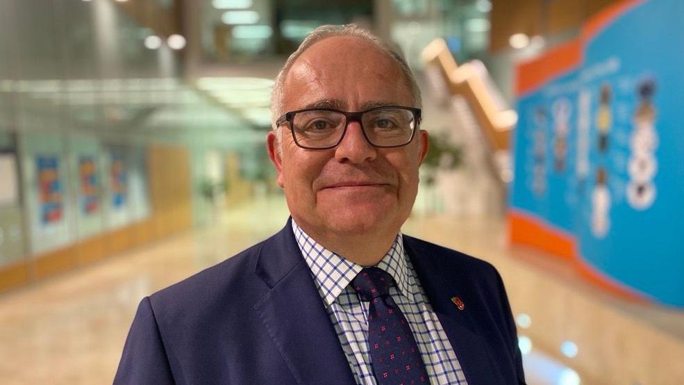 Paul Weston standing alone. He is wearing glasses and a navy suit with a poppy badge on his left lapel. He is wearing a checked pattern shirt and a tie. The background of the image is blurred