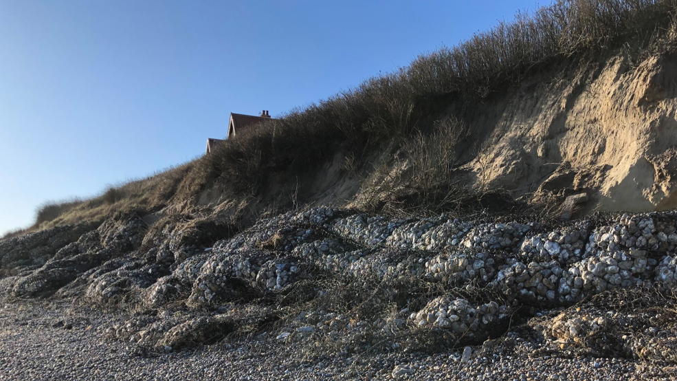 Geobags of rocks form sea defences at the foot of an eroded cliff on a gravel beach, with a house seen in the background behind long grass at the cliff edge.