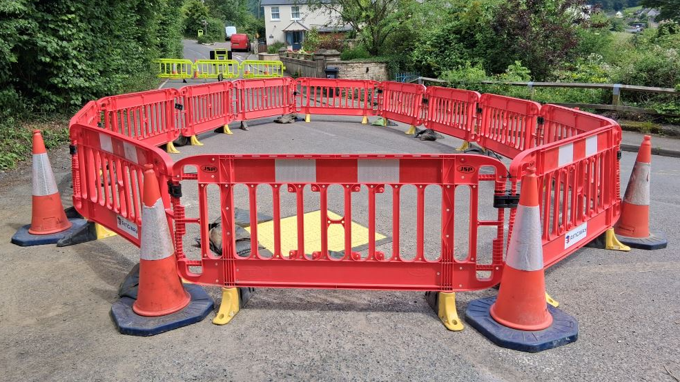 A large circular arrangement of cones and bollards blocks a quiet leafy road.
