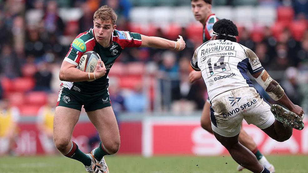 Jack van Portvliet stands with his feet together and his right arm wrapped around the ball with his left arm out straight behind him as Kaleveti Ravouvou runs towards him