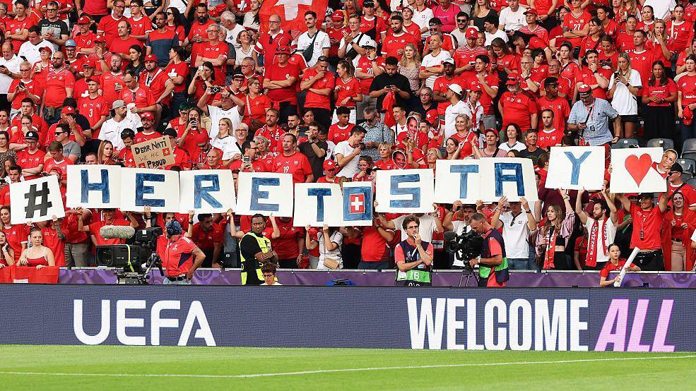 Fans of Switzerland display a TIFO reading "#heretostay"