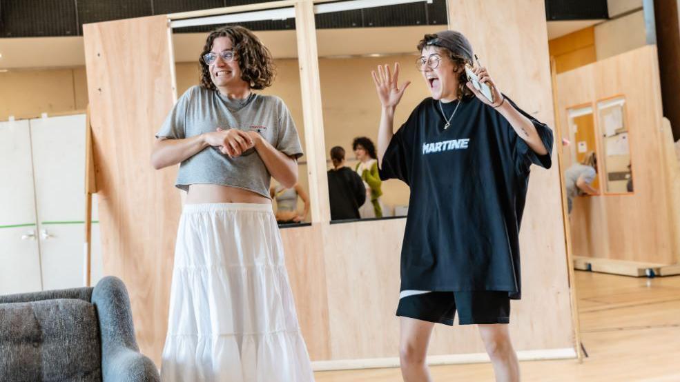 Two people stand smiling off camera. One wears glasses, a great shirt and white long skirt, while the other wears a cap, oversized black t-shirt and sports shorts