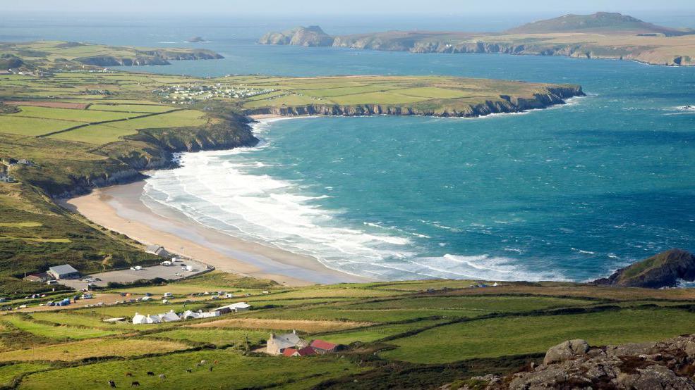 A look down on a sandy beach and clear blue sea. Green hills surround it, with cattle and farmhouses dotted about.