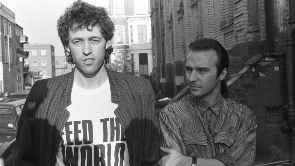 An archive shot of Bob Geldof wearing a T-shirt that says 'feed the world' standing next to Midge Ure.