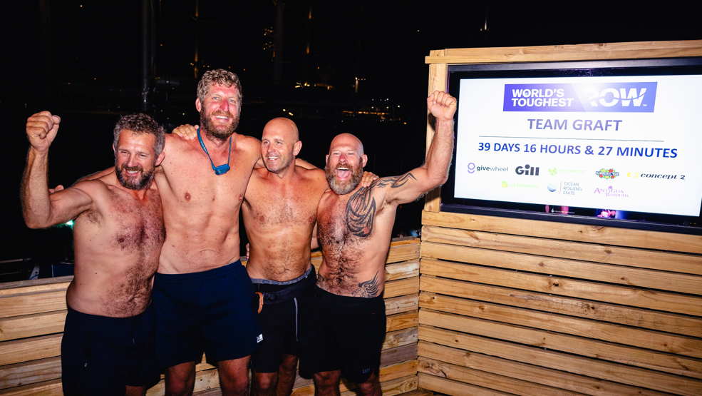 Four men are standing bare chested and wearing black shorts in front of a sign which gives their time as 39 days, 16 hours and 27 minutes.