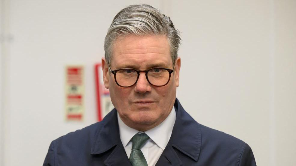 Picture of Keir Starmer - he has glasses on and a dark navy trench coat with a green tie.