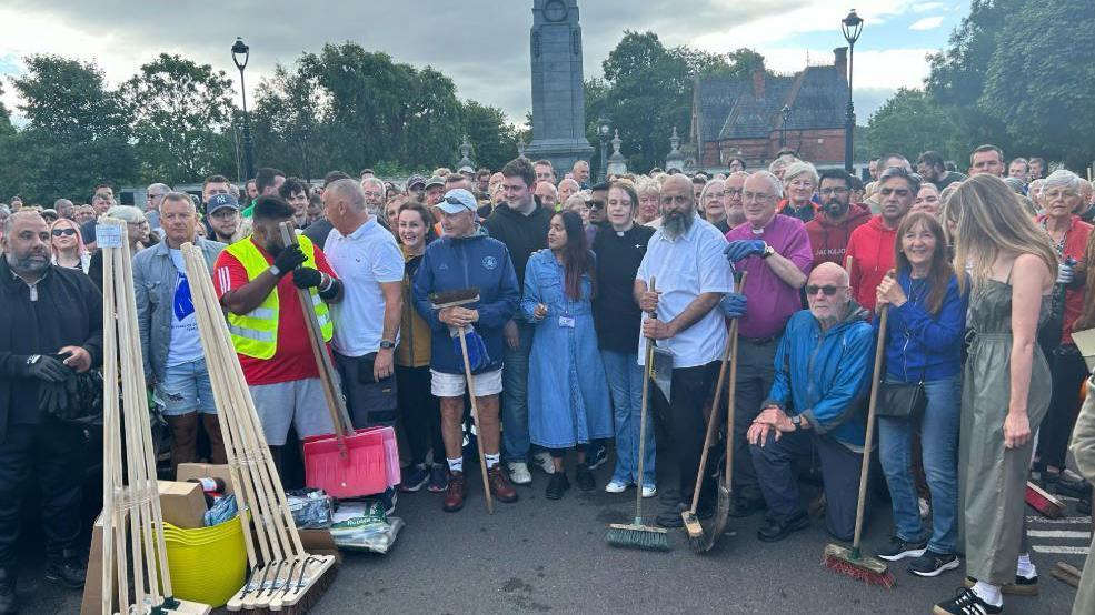 A crowd consisting of dozens of people, holding brooms and brushes, with a pile of brushes and buckets to the left. They are gathered in an open space with a cenotaph behind them.