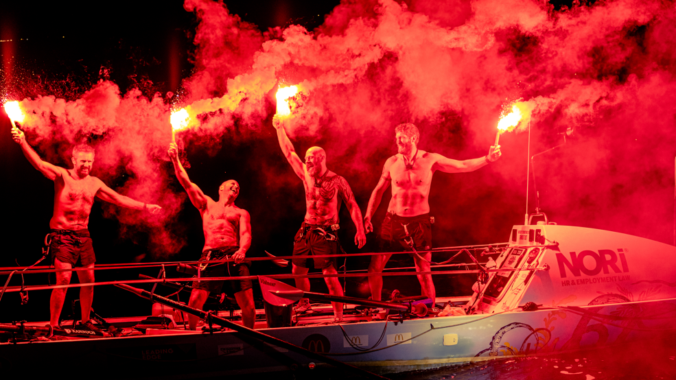 Four men in shorts  are standing on an open boat holding red flares. They are all topless and are cheering and laughing.