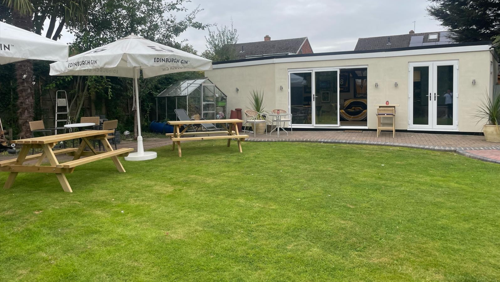 The white exterior of the bar can be seen with a small patio path right outside. There are umbrellas and pub benches in the garden to the left of the bar.