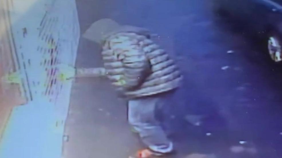 A man in a dark puffer jacket and hoodie sprays on a wall in a grainy colour shot taken from a CCTV camera above.