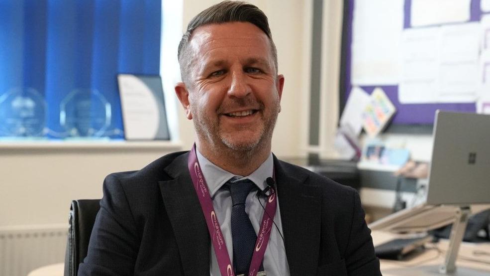 A man in a navy blue suit and tie and a blue shirt, sitting in an office with a purple lanyard. He is smiling.