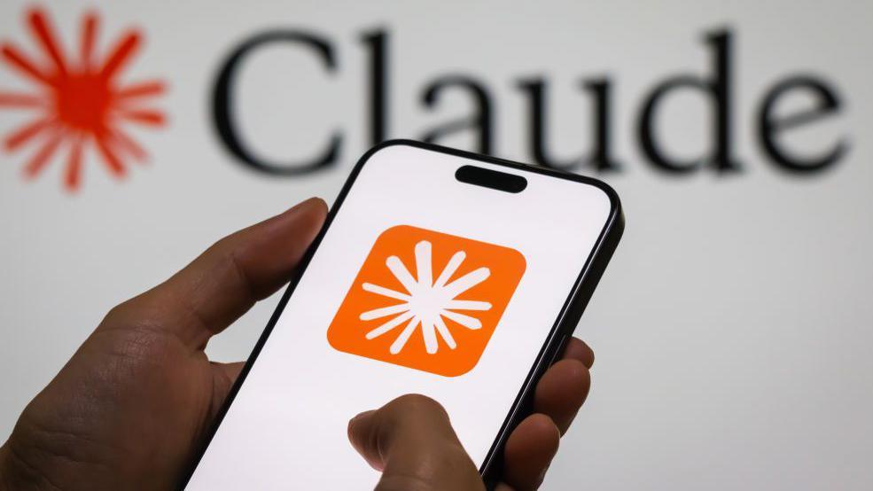 Close up of hands holding a mobile phone displaying an orange and white icon. On the wall behind it says Claude