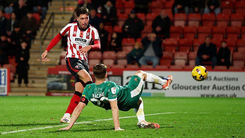 Isaac Hutchinson stands on one leg after striking the ball which flies through the air towards the goal, as a Notts County player crouches on the floor with a leg outstretched trying to block it