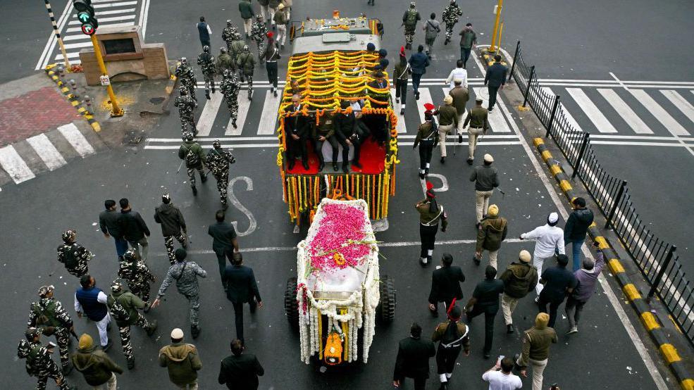 Funeral procession shows several people walking behind a coffin covered in colourful flowers on a street in New Delhi