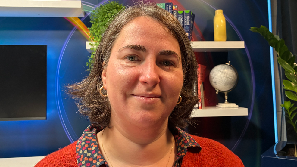 Dr Alexandra Tomescu smiles at the camera in an office. She has short brown hair and wears  earrings and a red sweater over a patterned collared shirt.