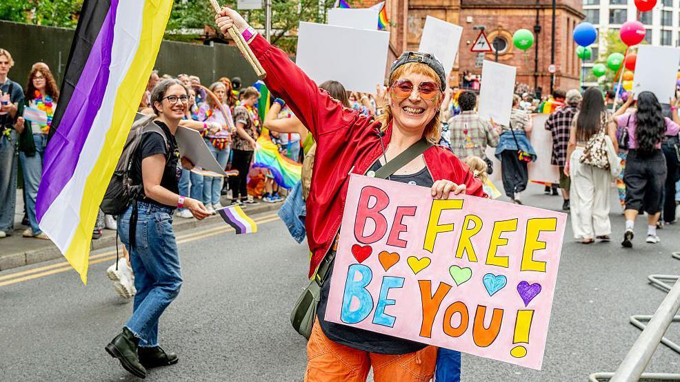 A photograph of part of Manchester Pride's parade in 2025. Someone is holding a colourful banner which says 'be free, be you' on it. They are wearing brightly coloured clothing including a red jacket, orange trousers and tinted sunglasses. They are waving a flag in their other hand. Crowds are gathered on a pavement and on the street behind them.