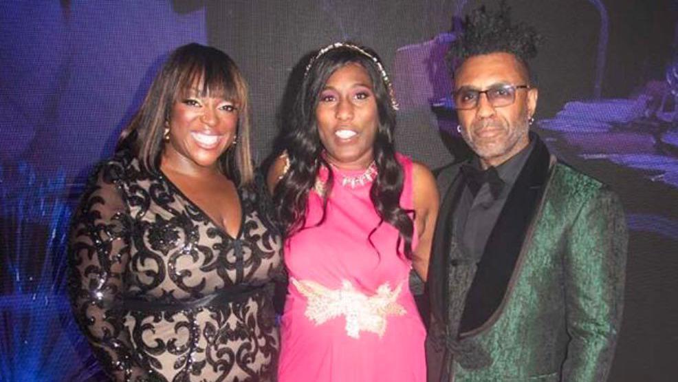 Two women and a man stood against a purple backdrop looking at the camera. The woman on the left is wearing a black and beige dress and the woman in the middle is wearing a pink dress. The man, stood on the right, is wearing a green tuxedo