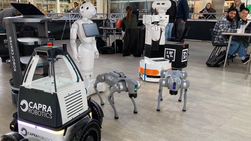 Some robots are in a shopping centre. There is one that looks humanoid and two that look a little like dogs. People are sitting at tables close by, and some people are looking at the robots from the side of the area