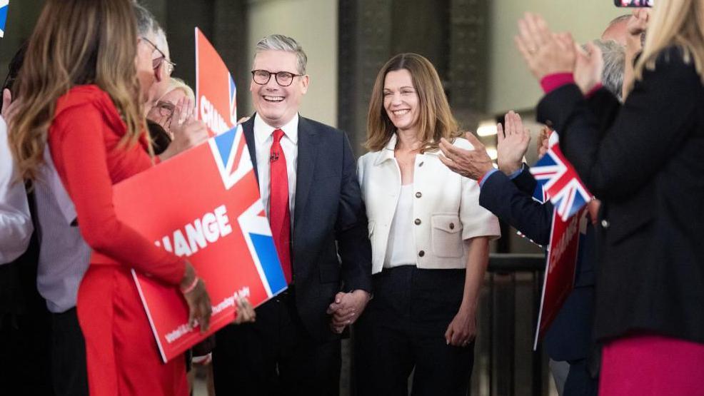 Keir Starmer and his wife Victoria smile as they walk through clapping crowds.