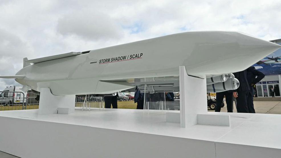 A Storm Shadow cruise missile, which is several metres long and has a white glossy outer layer, as well as a "Storm Shadow/SCALP" logo, pictured on a display stand at an air show in Farnborough 