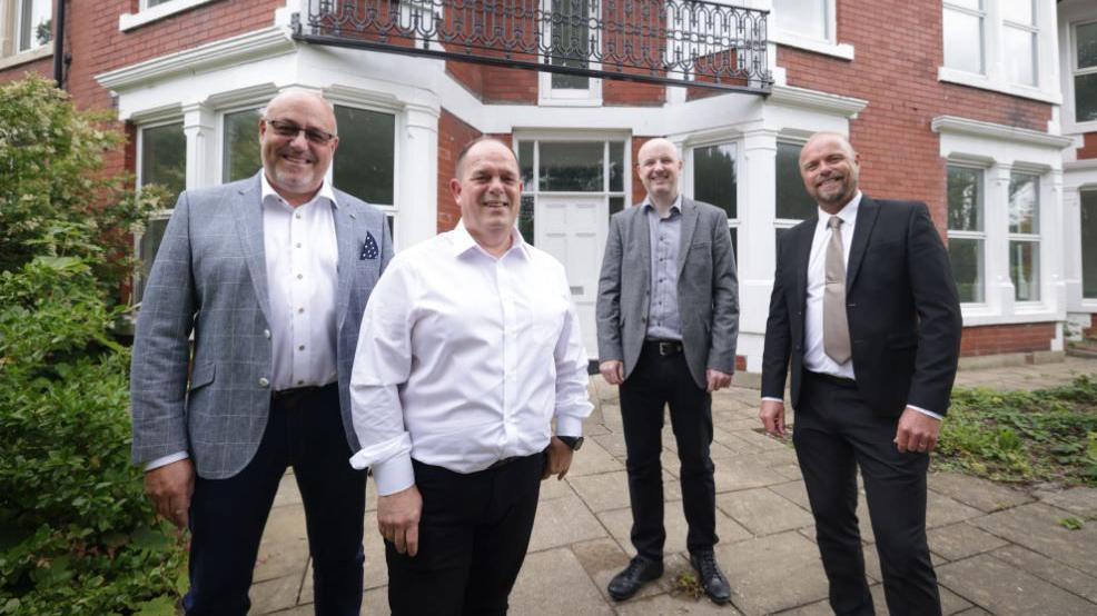 Four men involved in the project outside the large former care home. It is redbrick and its columns are painted in white.