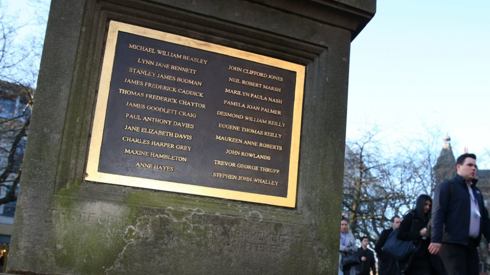 A stone memorial column with a large gold edged brown plaque showing 21 names written in gold. Pedestrians can be seen walking past