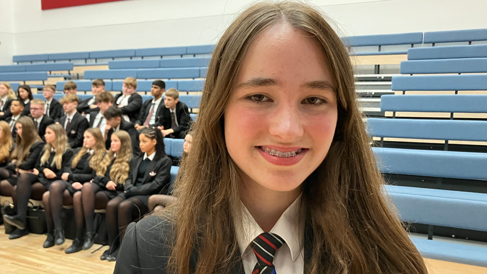 A girl wearing a blouse, tie and grey jacket in a school hall with a number of other children sat on benches behind her