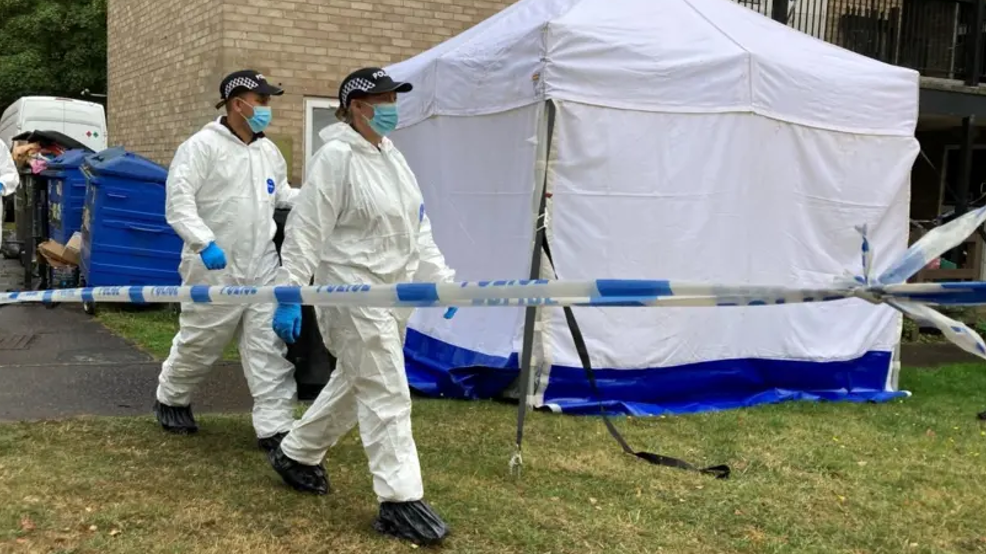 Two police officers in white forensics suits walk past a white police tent. There is a police cordon in front of the tent.