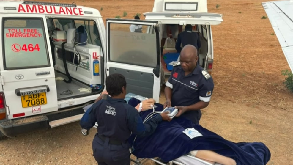 A man covered in a blue blanket on a medical gurney, there are two medics standing either side of him wearing blue overalls. Next to him is a white ambulance with an open door