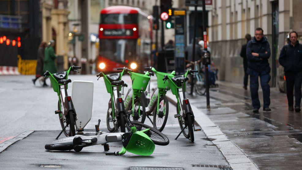 A group of lime coloured bikes on a street in central London. A bus is visible in the background. One of the bikes has fallen over.