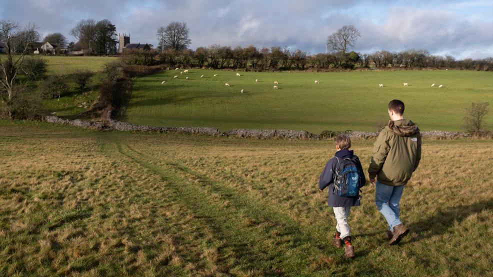 A boy and a young male adult walk through a field in Priddy, Somerset, wearing walking boots and coats. In the background there is a field of sheep, a church and stone walls. 