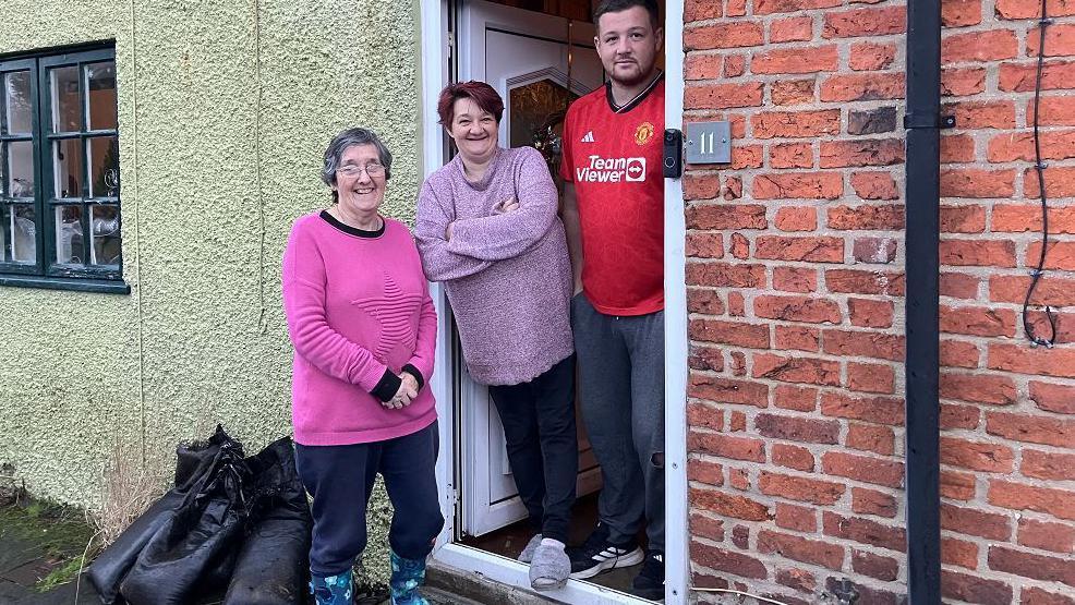 Sylvia Fisher, a woman in a pink jumper and blue trousers and wellies, with a couple at the door of a terraced house which has flooded 