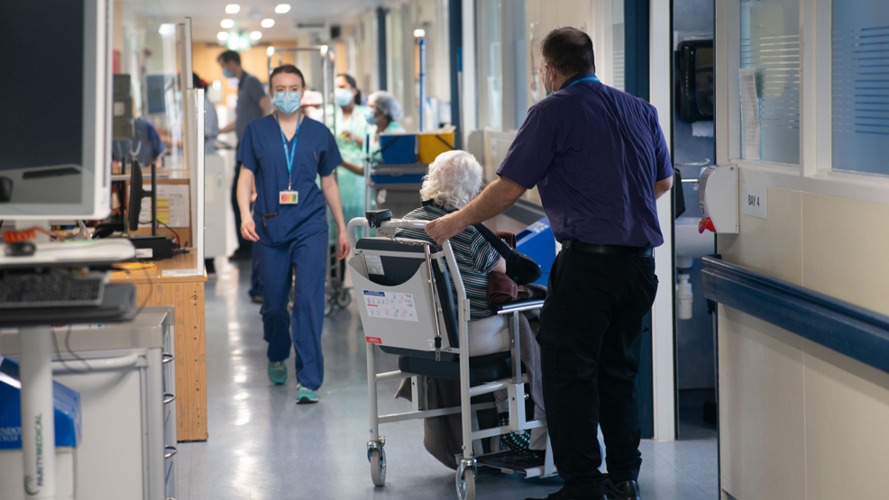 A stock image of a busy hospital. A doctor in blue scrubs walks towards the camera, while and elderly person is supported in a wheelchair. 