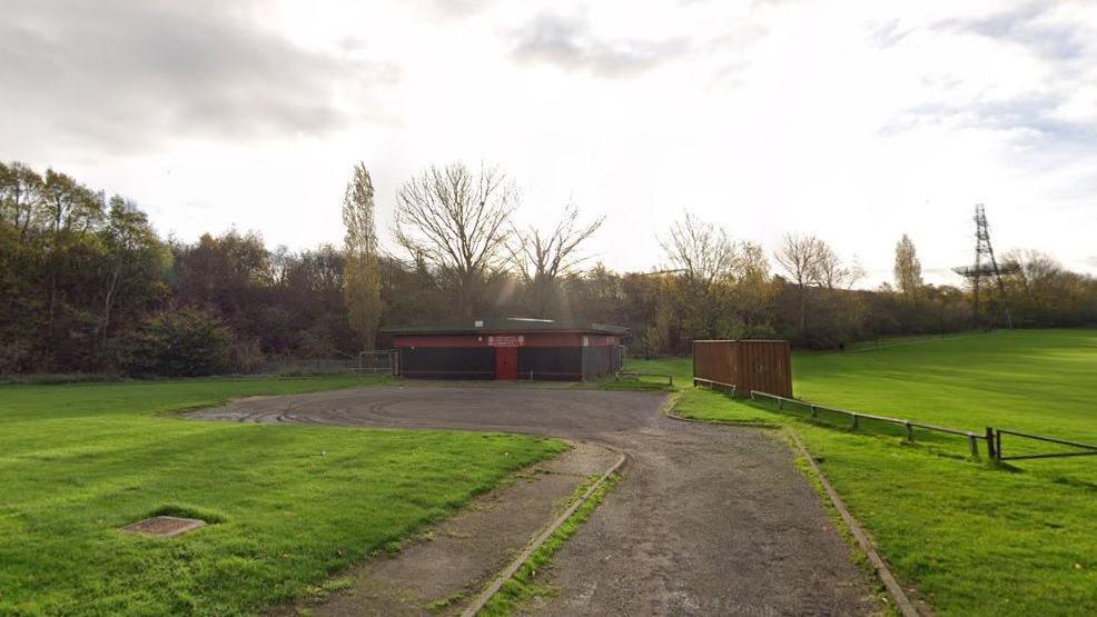 Red and black single-storey club house with car park in front. A playing field with a small fence is visible to the right. There is a narrow road leading to the car park, with a footpath and grass to the left