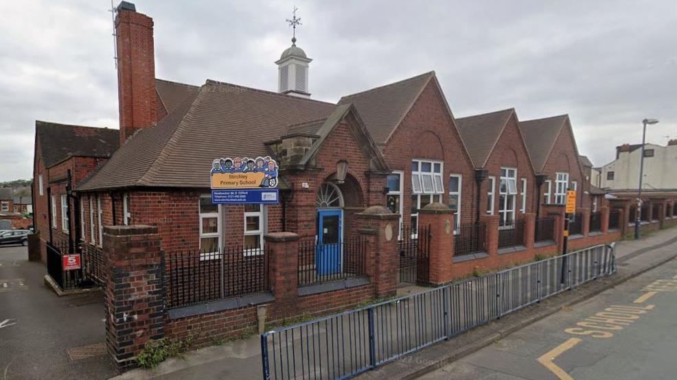 Stirchley Primary School is a single-storey red brick building with pointed roofs and a blue door. A blue and yellow sign is outside the entrance. There are railings along the pavement and yellow road markings. The school windows are open.