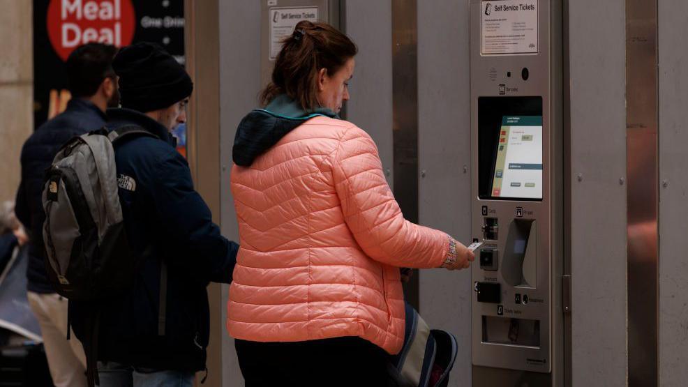 Woman wearing a pink puffa jacket buys a train ticket from a self-service machine using a debit or credit card