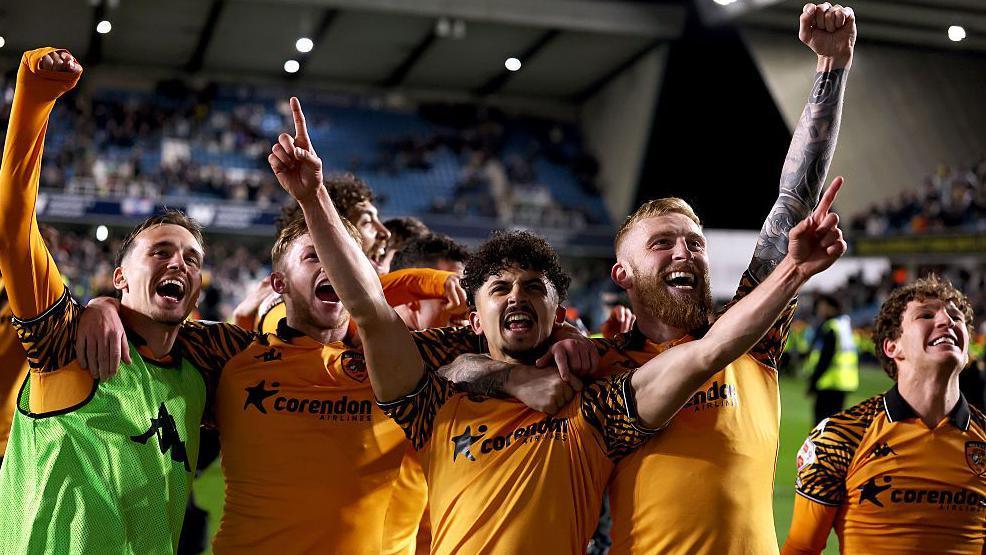 Mohamed Belloumi and Oliver McBurnie of Hull City celebrate at full time following the team's victory in the Championship play off semi-final second leg match against Millwall. They are joined by other players and all are wearing orange football shirts.