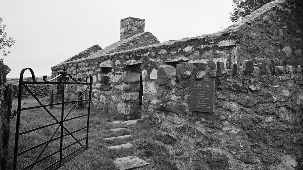 A black and white image of a stone cottage, with a black gate in the foreground